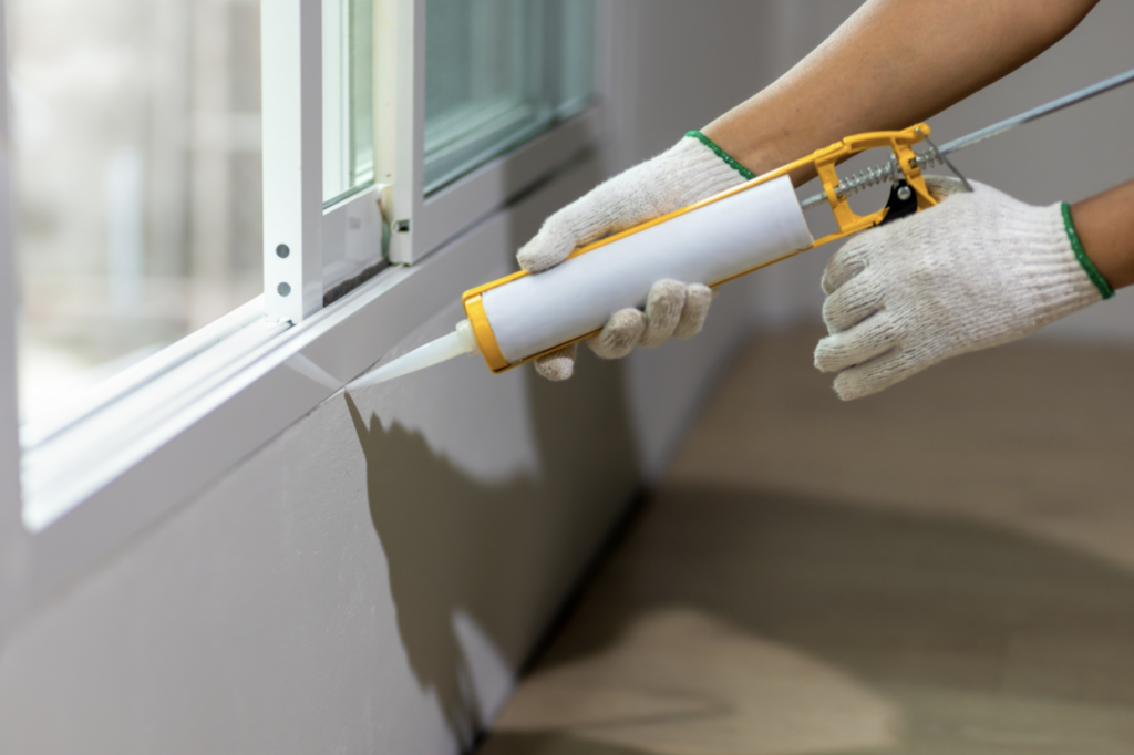 person wearing gloves uses caulk gun with caulk to seal gap between window and wall on inside of home